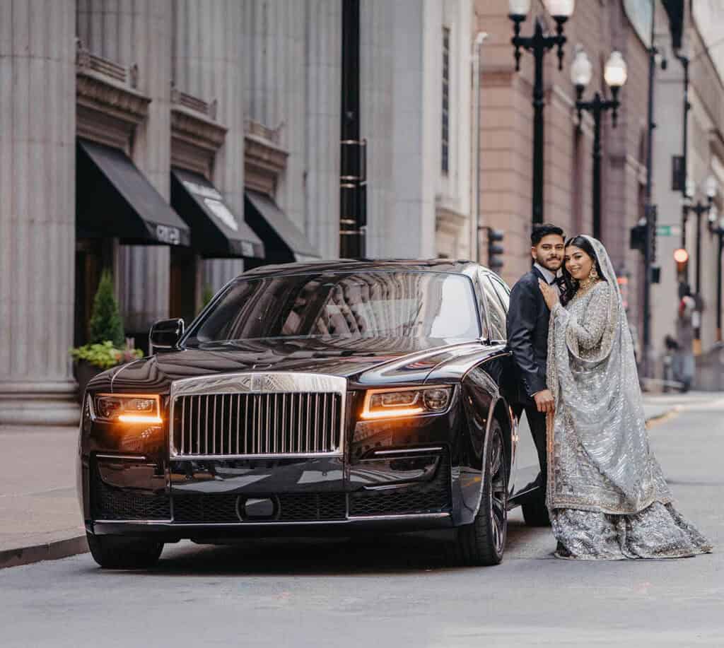 Luxury wedding couple in elegant attire with chauffeur-driven black limousine in city street.
