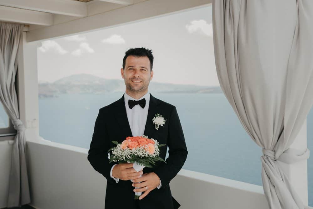 Elegant groom in a black tuxedo holding a floral bouquet at a luxury seaside wedding venue.