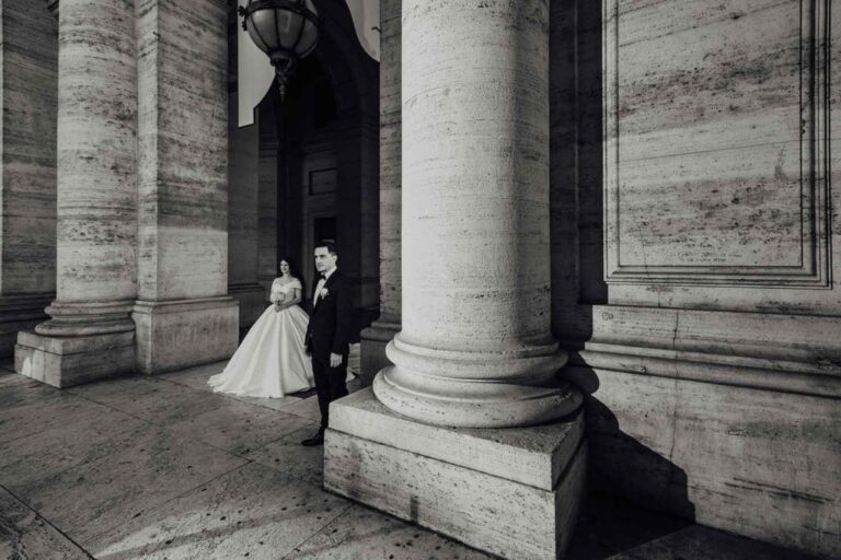 Elegant bride and groom standing outside a grand classical wedding venue with tall stone columns and dramatic architecture.