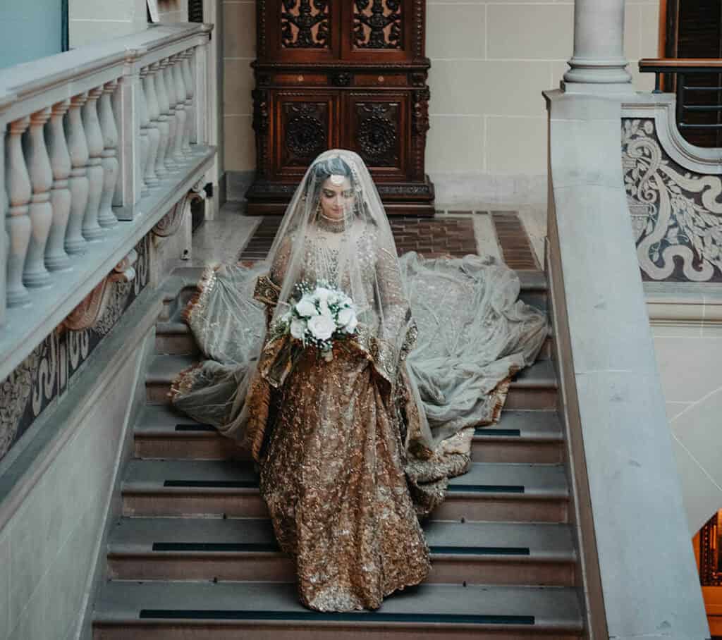 Elegant bride in ornate traditional wedding dress with veil holding a bouquet on grand staircase at luxury venue.