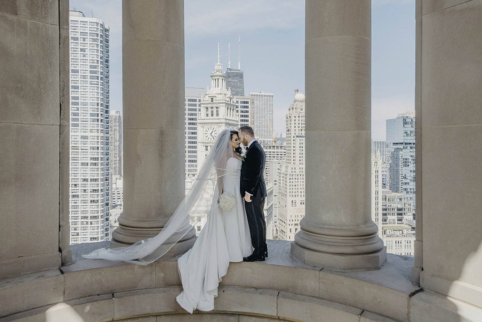 Elegant wedding couple stands atop a historic building with panoramic city views of Chicago's skyline, showcasing luxury wedding photography.