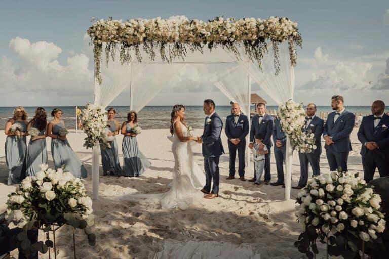 Luxurious beach wedding ceremony under a floral canopy on sandy shores, with elegant bridal party and ocean backdrop.