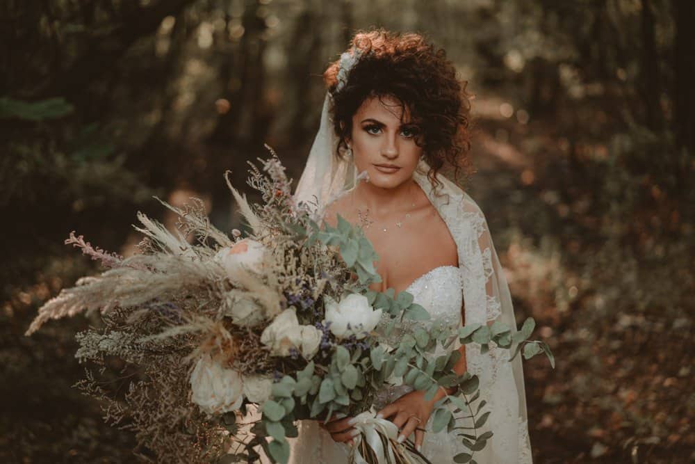 Vow bride holding a large bouquet of wildflowers and greenery in a lush forest setting, perfect for a romantic wedding photoshoot.