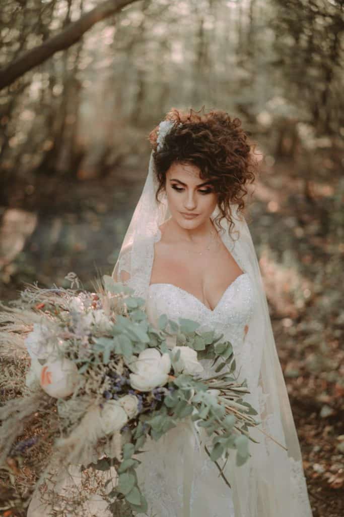 Bride with curly hair holding a lush floral bouquet in a forest setting, perfect for luxury wedding photography.
