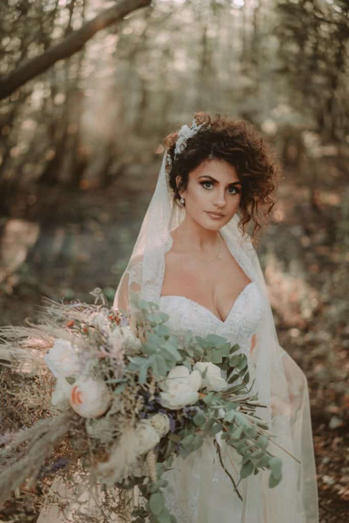 Elegant bride with floral bouquet in a forest setting for luxury wedding photography.