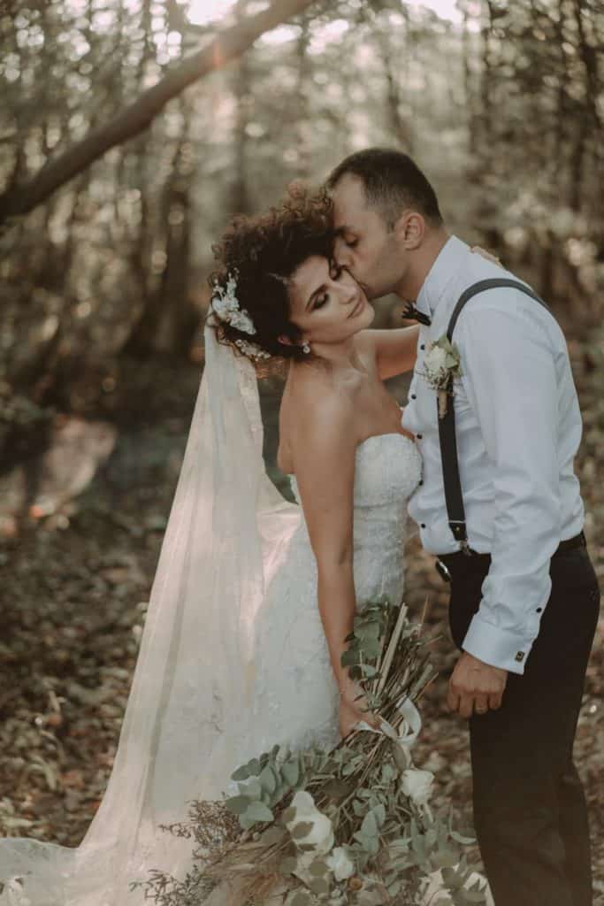 Elegant outdoor wedding photo of a bride and groom sharing a kiss in a forest setting, capturing romantic and high-end wedding vibes.