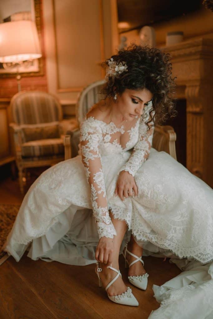 Elegant bride in lace wedding dress putting on heels in luxurious vintage room.