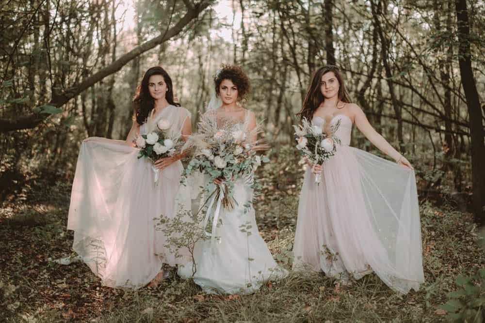 Elegant bridal models in flowing white dresses holding floral bouquets in a lush woodland setting for a luxurious wedding photoshoot.