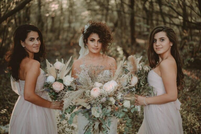 Elegant bride with floral bouquet surrounded by bridesmaids in a lush outdoor setting for a luxury wedding.