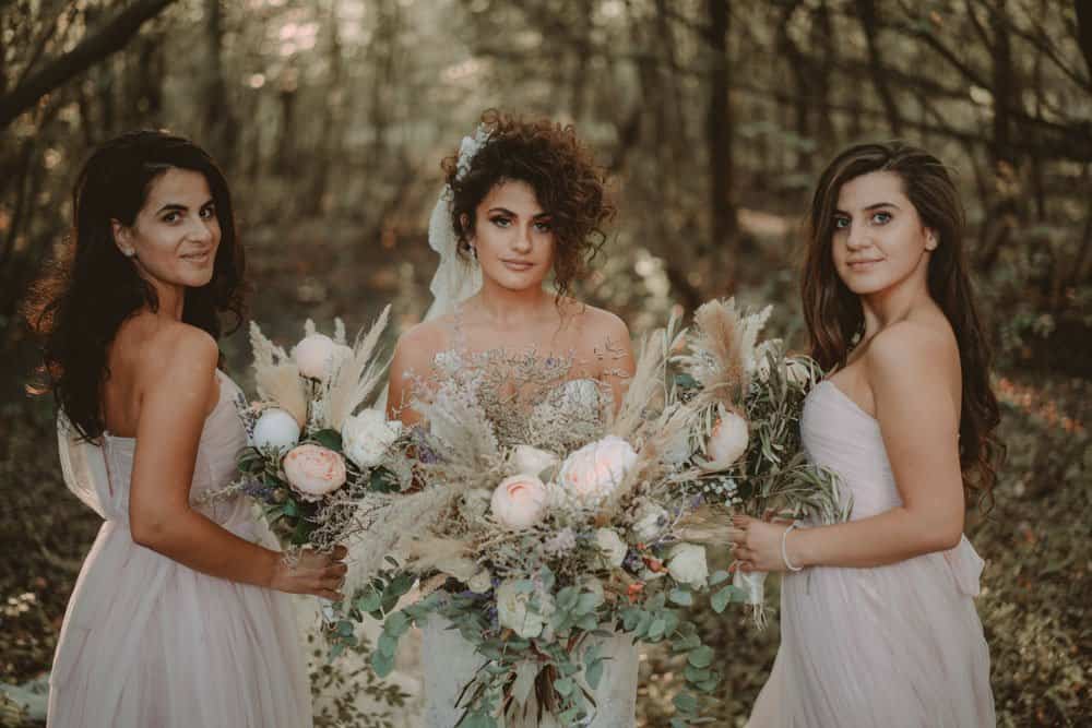 Elegant bride with floral bouquet surrounded by bridesmaids in a lush outdoor setting for a luxury wedding.