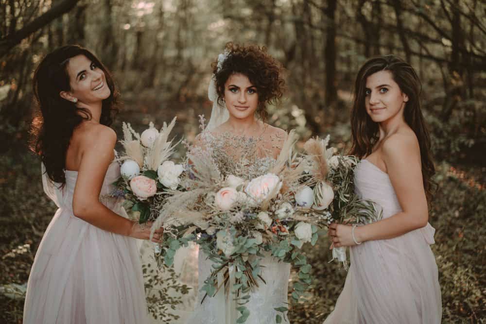 Beautiful bride with two bridesmaids holding wedding bouquets in a romantic outdoor woodland setting, perfect for luxury wedding photography.