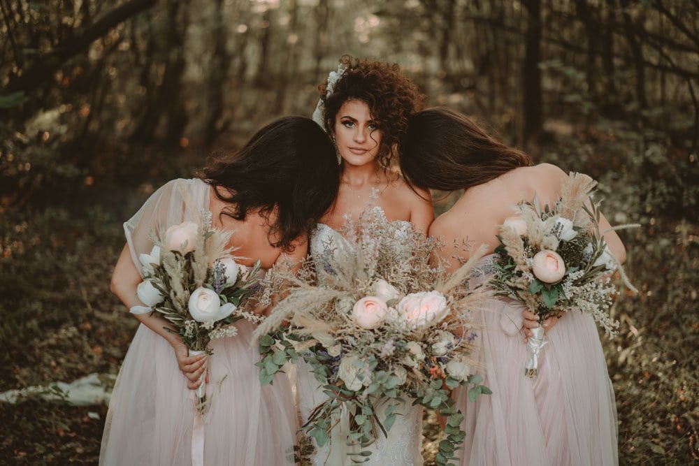 Elegant bridal portrait with three brides holding lush flower bouquets in a woodland setting, showcasing a romantic outdoor wedding theme.