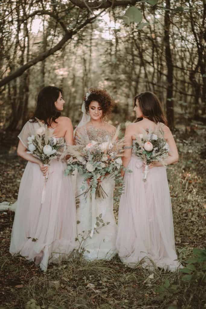 Elegant wedding bridesmaids in flowy pastel dresses holding floral bouquets in a forest setting.
