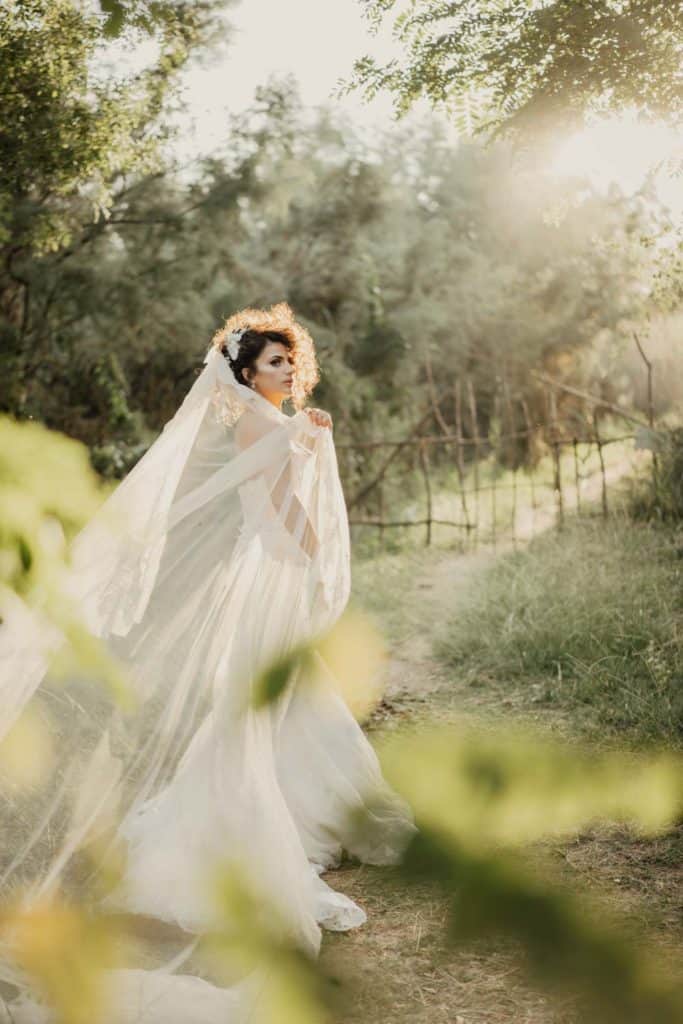 Elegant bride in flowing white gown with veil, in lush outdoor setting at sunset.