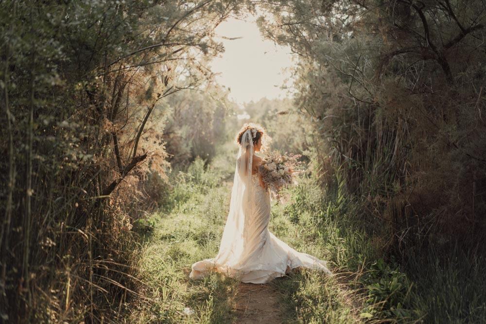 Elegant bride in a flowing white wedding gown holding a bouquet in a nature-filled outdoor setting with warm, natural sunlight.