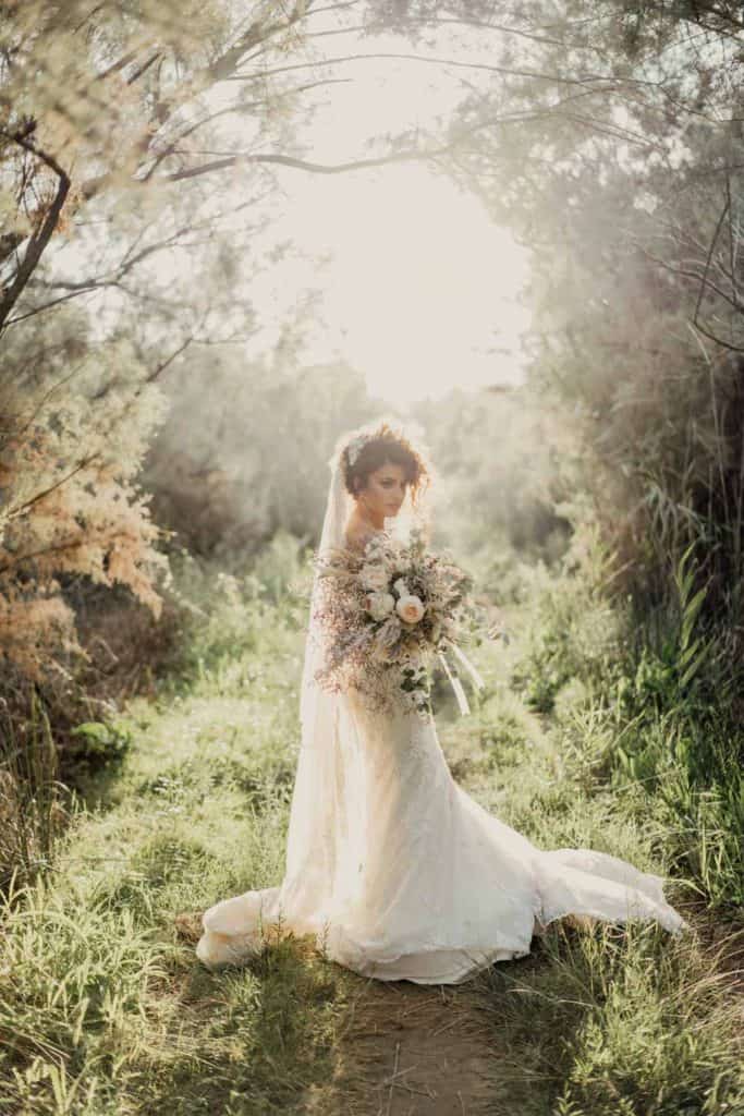 Elegant bride holding a floral bouquet in a serene outdoor setting with soft sunlight.