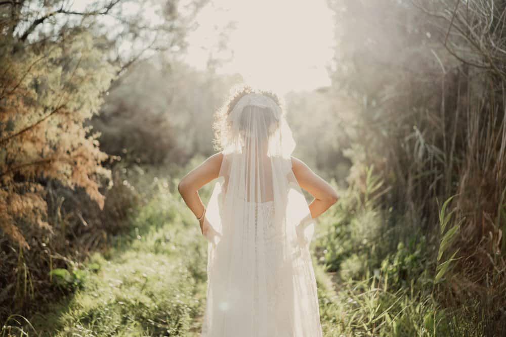 Elegant bride standing in a lush outdoor setting during sunset, showcasing a luxurious wedding gown and veil.