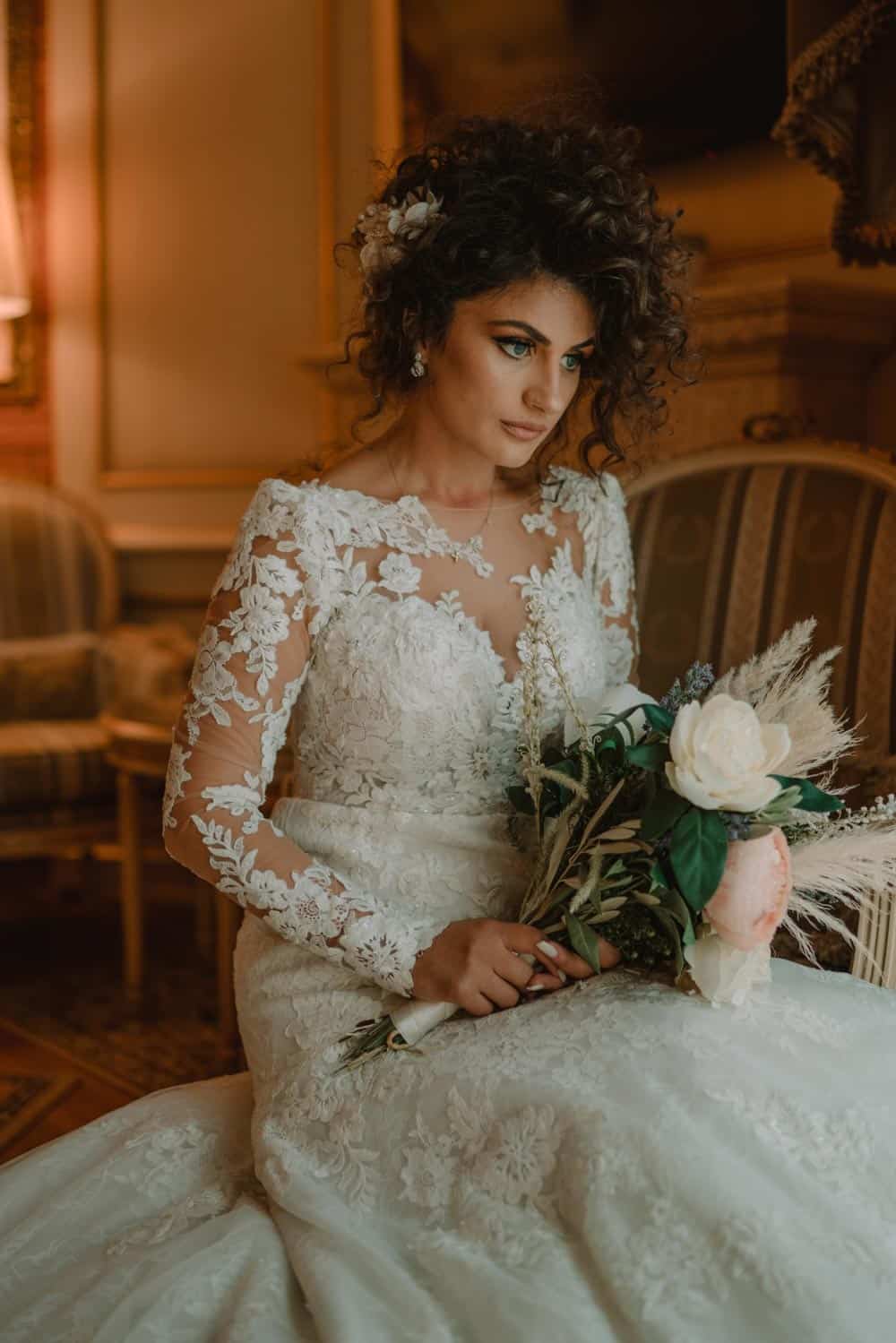 Luxury bride in lace wedding dress holding bouquet in glamorous interior setting.