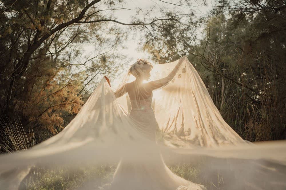 Elegant bride in flowing gown with veil in a luxurious outdoor wedding setting at sunset.