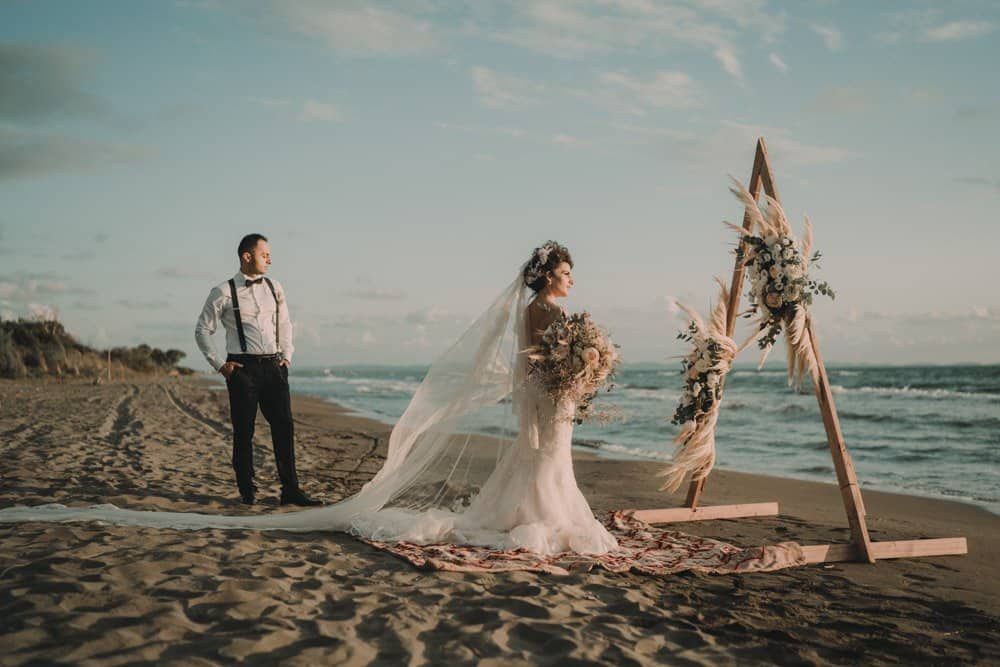 Beach wedding ceremony with bride and groom by the ocean, elegant floral arch, luxury wedding setup on sandy shore, high-end destination wedding in a scenic coastal environment.