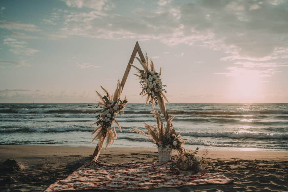 Elegant beach wedding arch with floral arrangements, set against a serene sunset by the ocean. Perfect for luxury seaside wedding ceremonies.