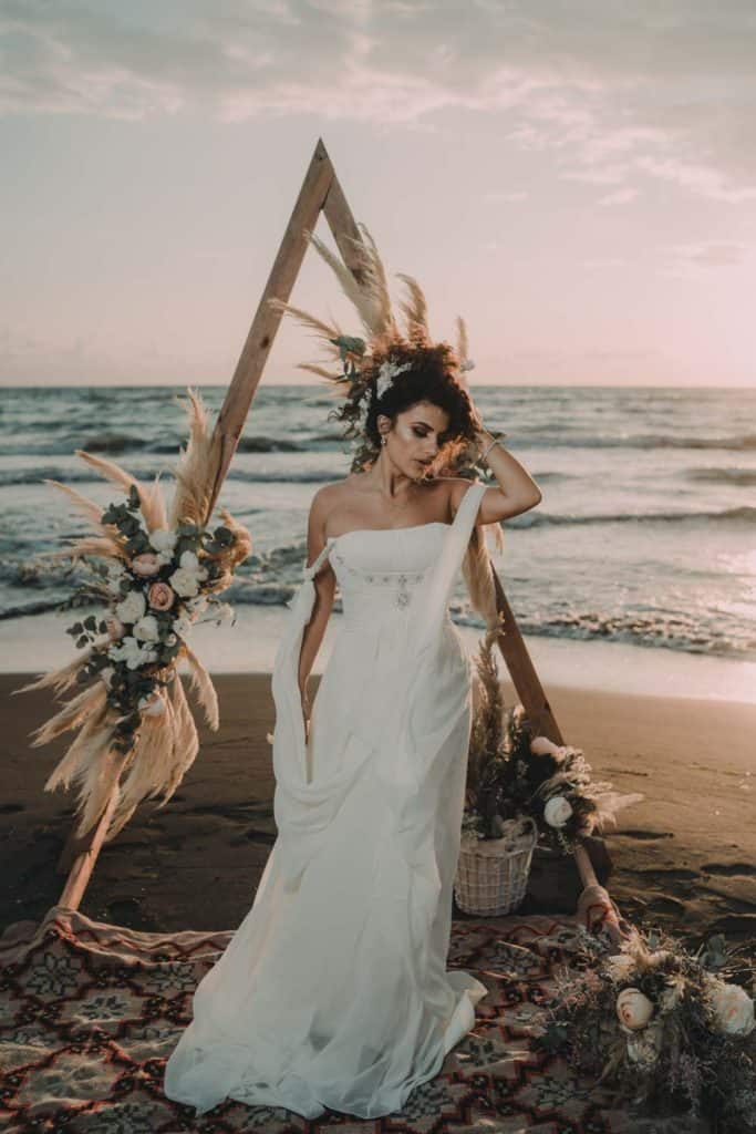 Elegant bride in a flowing white wedding gown on the beach with boho floral arch during sunset, perfect for luxury destination wedding photography.