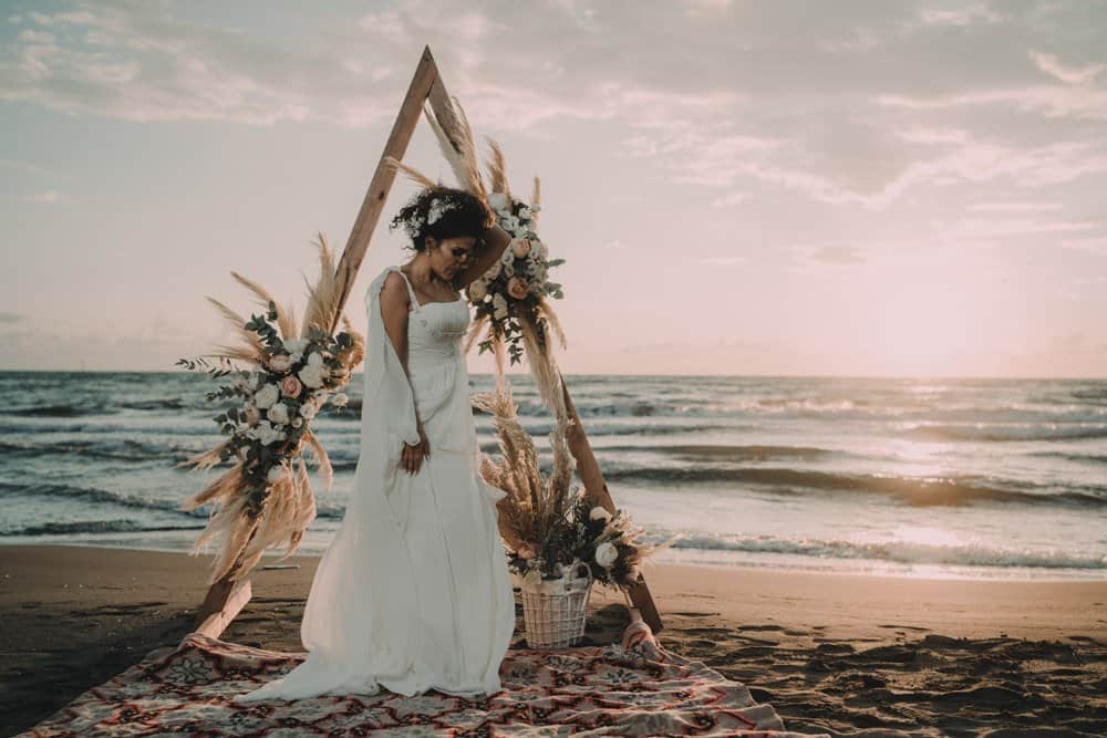 Elegant bride in a white wedding gown standing on a beach at sunset, showcasing a luxurious wedding setting by the ocean.