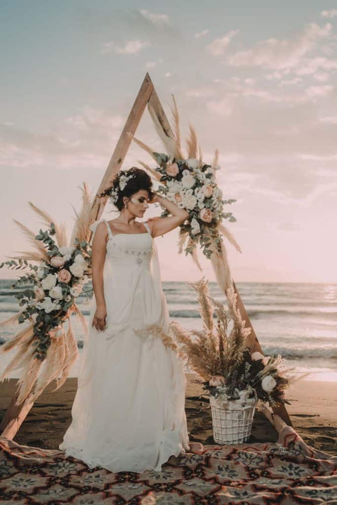 Elegant bride standing at a luxurious beach wedding arch with floral arrangements during sunset, showcasing high-end wedding venue decor.