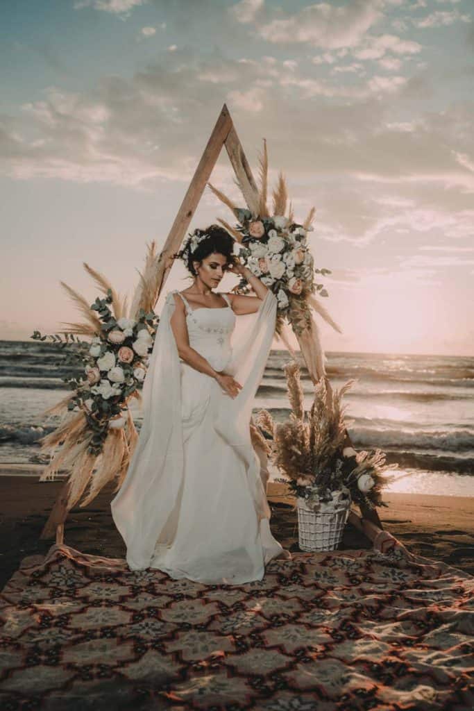 Elegant bride in a flowing white wedding gown standing on a sandy beach at sunset, decorated with luxurious floral arrangements and boho-chic decor.