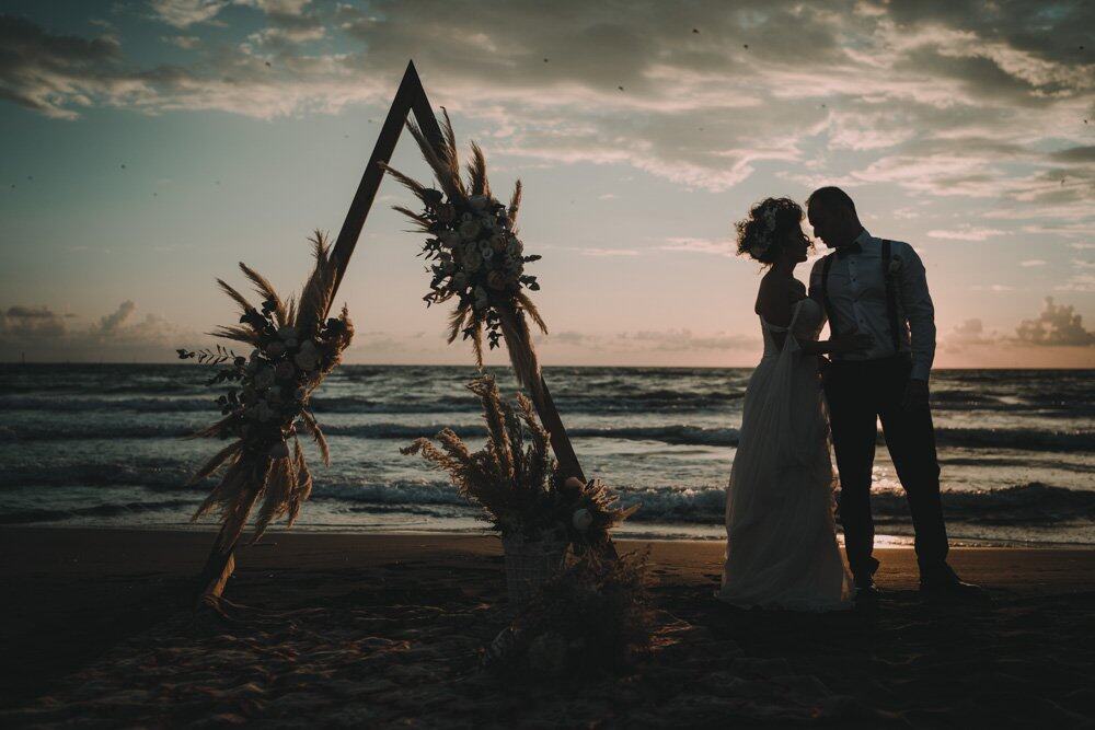 Elegant couple exchanging vows on a beach at sunset with a stylish wedding arch decorated with flowers. Perfect setting for a luxury destination wedding by Visistudio.
