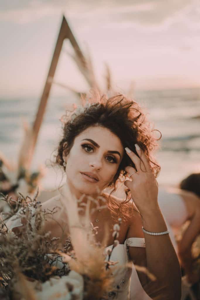 Elegant bride with natural makeup and curly hair at luxury beach wedding, scenic ocean backdrop.