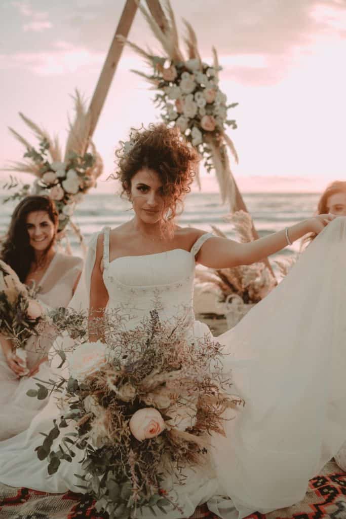 Elegant bride holding bouquet at luxury beach wedding during sunset, with high-end venue decor in the background.