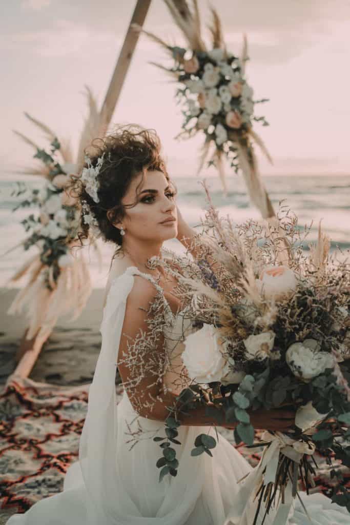 Bohemian bride holding a large floral bouquet at a coastal wedding decorated with a rustic arch and pampas grass.