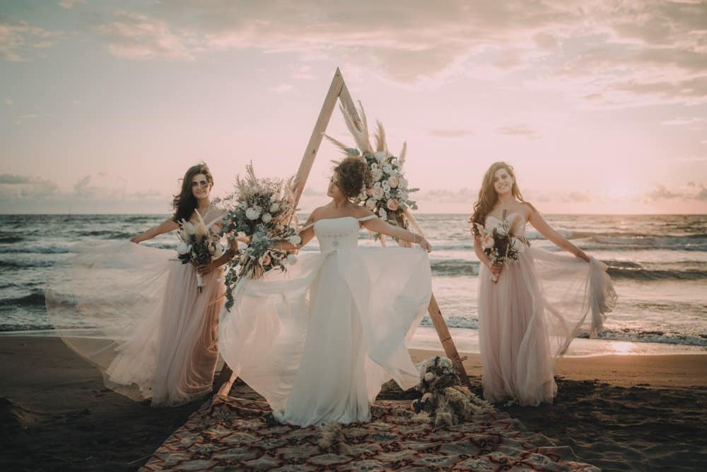 Elegant beach wedding with brides in flowing gowns and floral displays at sunset.