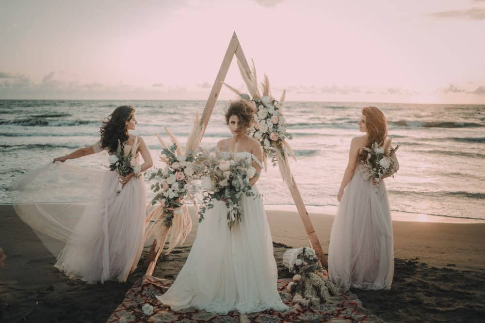 Elegant beach wedding with floral arch and bridesmaids at sunset, showcasing luxury wedding decor on the shoreline.
