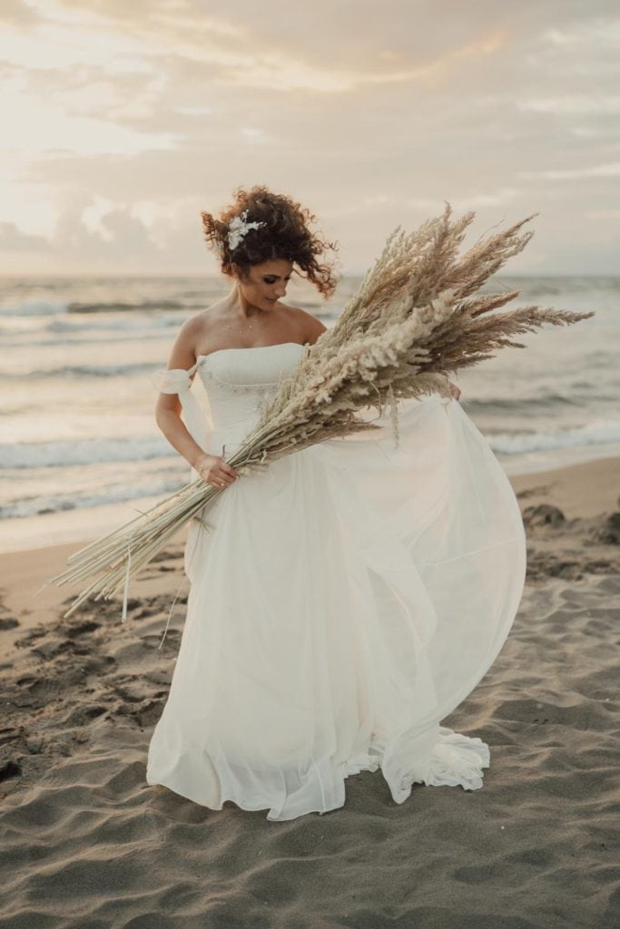 Gorgeous bride in a flowing white wedding dress holding pampas grass on the beach during sunset.