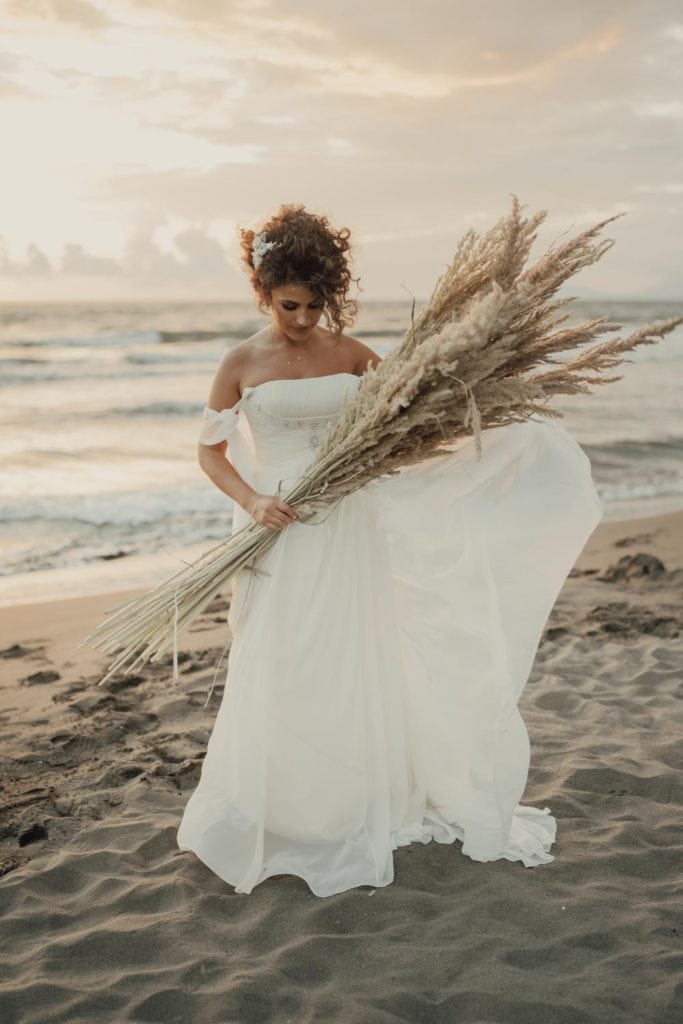Elegant bride holding pampas grass on the beach during sunset, perfect for luxury beach weddings.