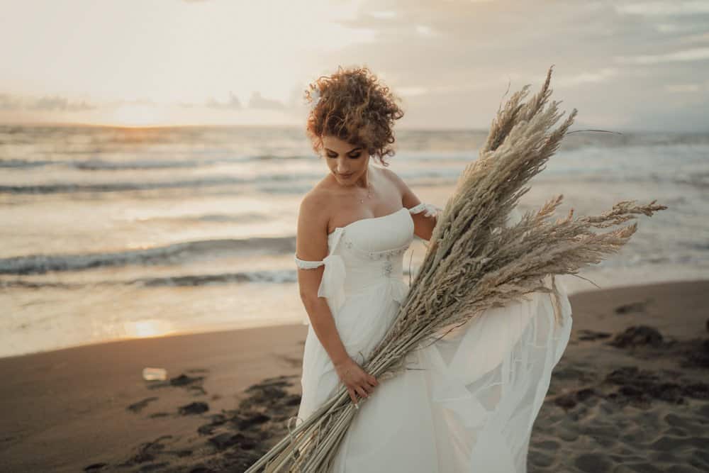 Elegant bride holding pampas grass on the beach during sunset, showcasing luxury wedding photography at a high-end seaside venue.