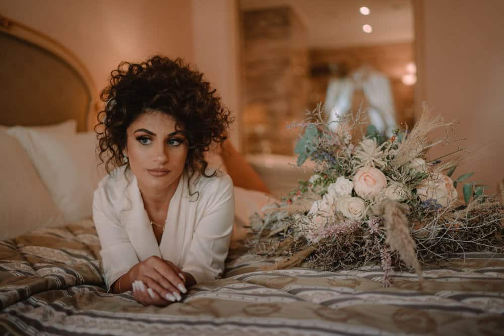 Luxury bride relaxing on plush bed with elegant floral bouquet in upscale wedding suite.