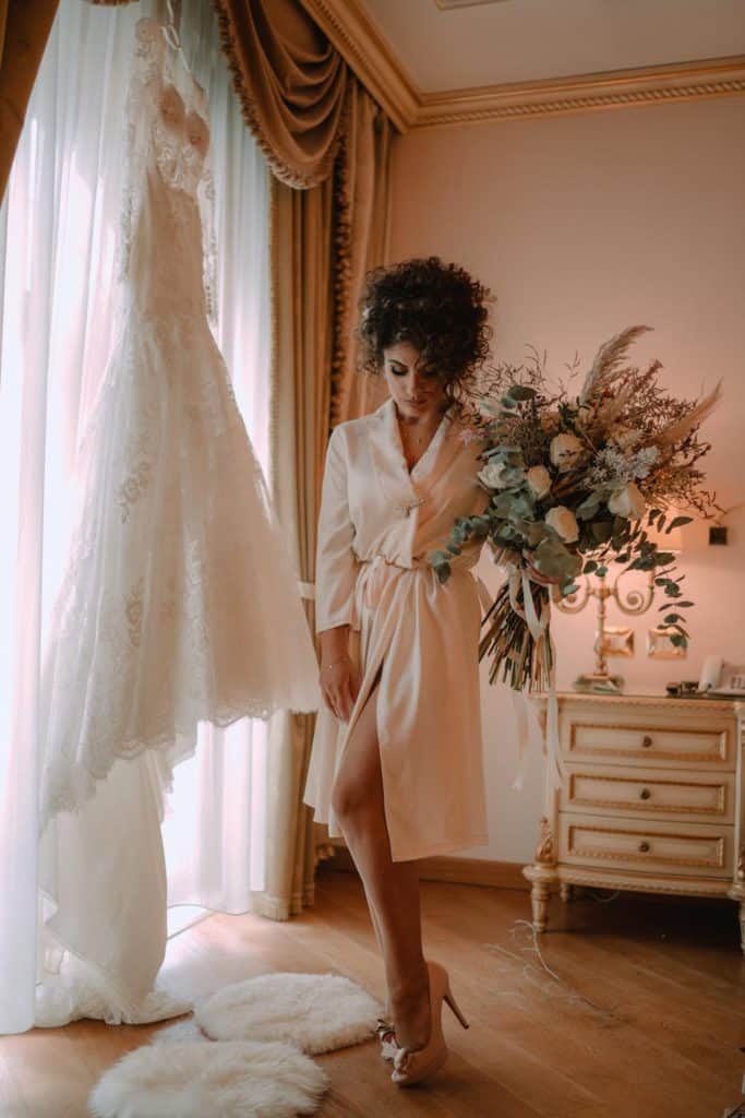 Elegant bride preparing for her wedding in luxurious bedroom, wearing silk robe, with wedding gown hanging nearby.