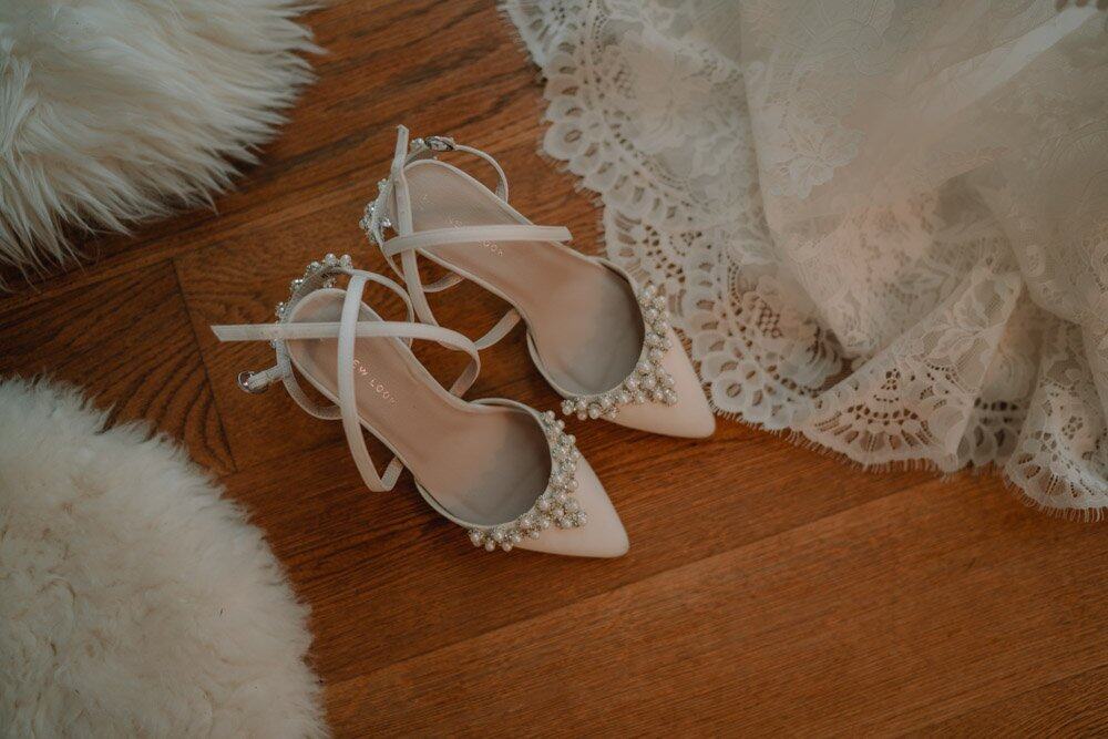 Elegant white wedding pumps with pearl embellishments on a wooden floor, surrounded by lace fabric and plush textures. Perfect for a luxury wedding setting.