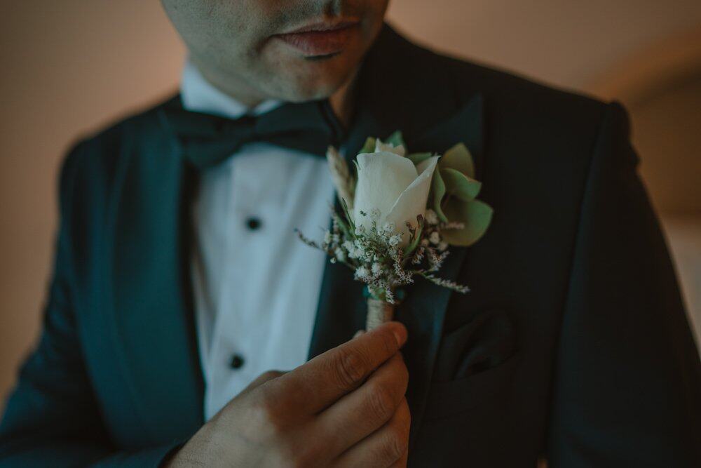 Elegant groom adjusting boutonniere of white roses and greenery, showcasing luxury wedding details.
