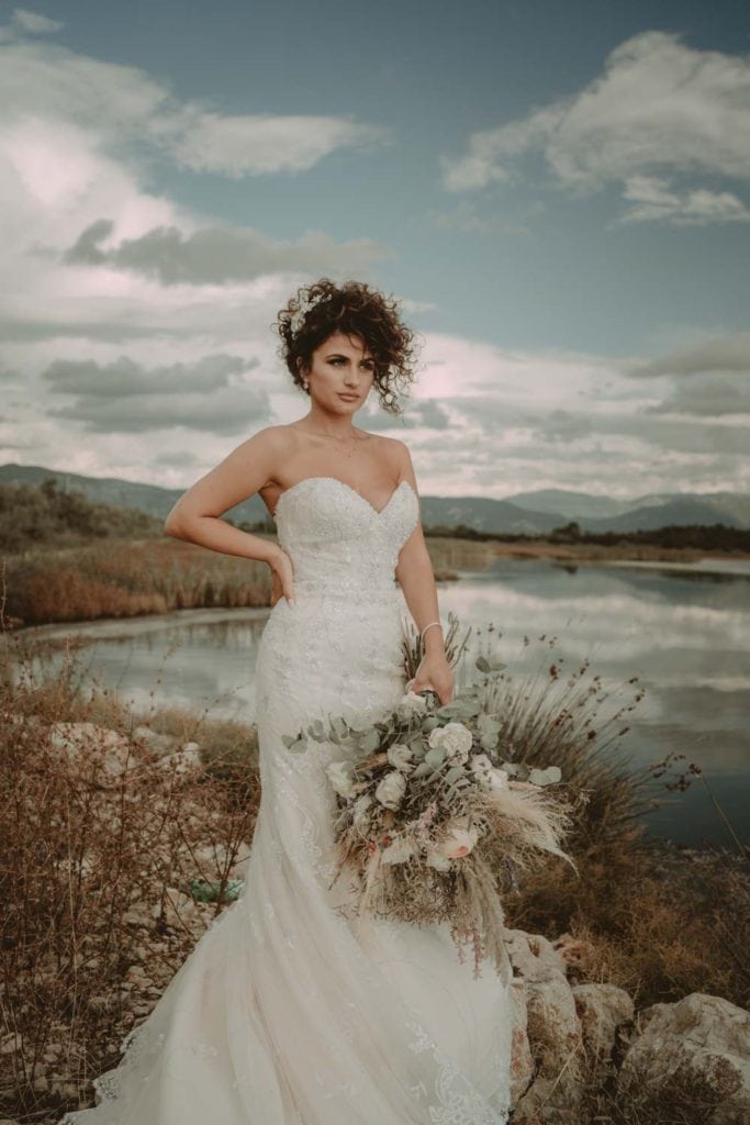 Elegant bride in stunning lace wedding gown holding boho floral bouquet in scenic outdoor setting.