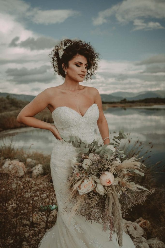 Elegant bride in a luxurious wedding gown holding a stunning bouquet at a scenic outdoor venue.