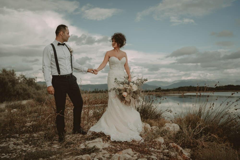 Elegant wedding couple holding hands outdoors by the water with scenic mountain views.