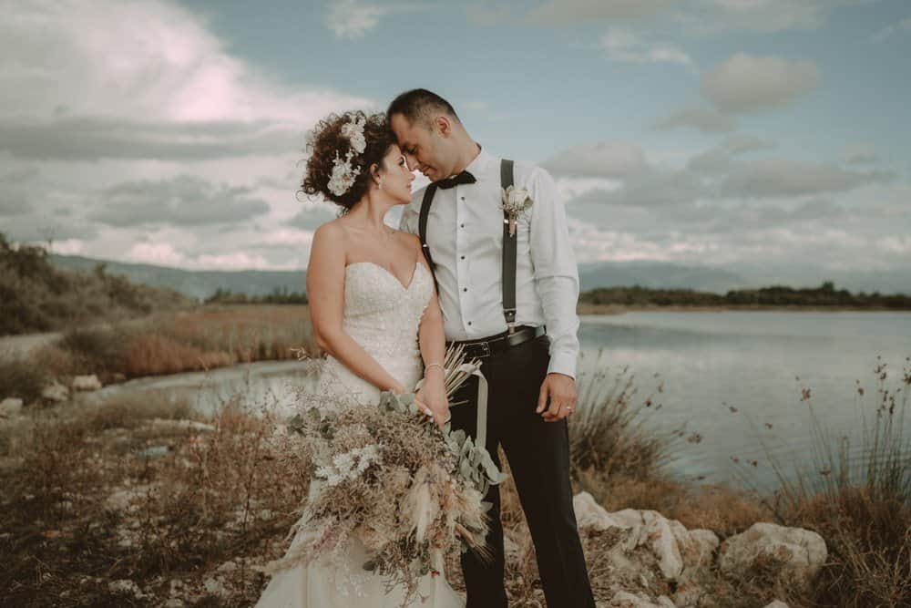 Elegant wedding couple by the lake in a scenic outdoor setting with cloudy sky and natural landscape.