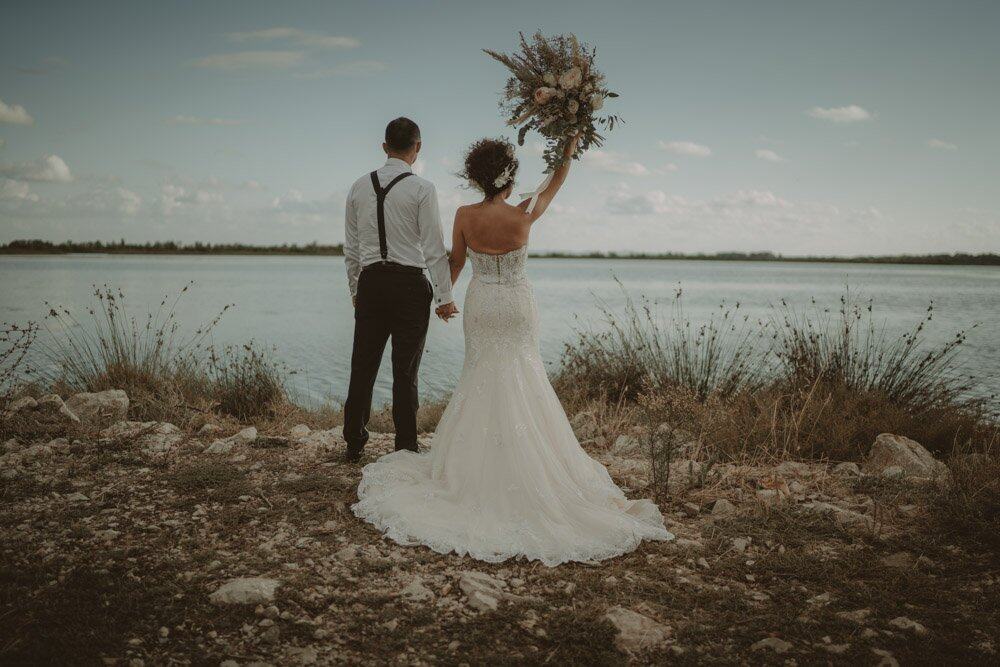 Elegant bride and groom holding hands by a scenic water body during a luxurious outdoor wedding.
