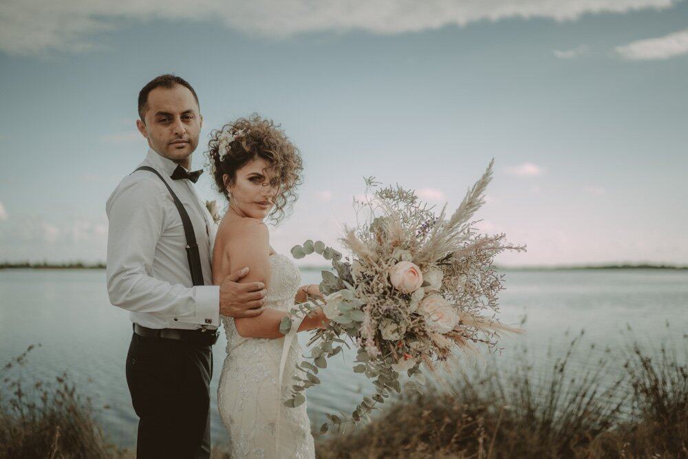 Elegant wedding couple by the water with a lush bouquet, showcasing luxury romantic wedding photography.