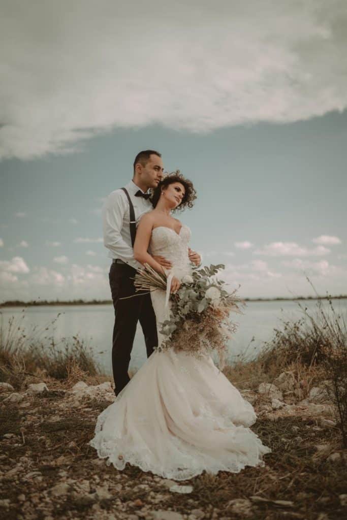 Luxurious wedding couple standing by the water with elegant attire and scenic outdoor backdrop, perfect for high-end wedding venues.