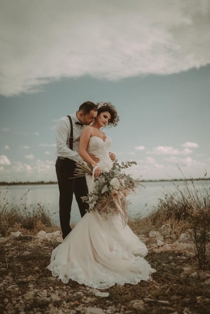 Elegant wedding couple outdoor photoshoot by the water, showcasing luxury wedding attire and romantic scenery.